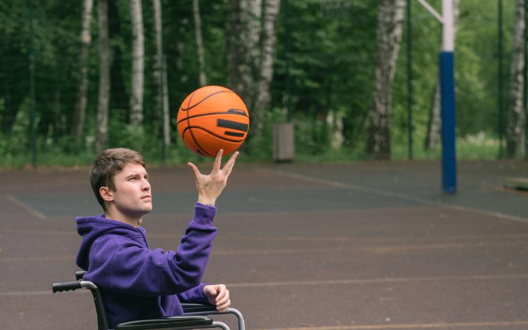 A young man in a wheelchair skillfully spins a basketball on an outdoor court. Un jeune homme en fauteuil roulant fait tourner adroitement un ballon de basket sur un terrain extérieur.