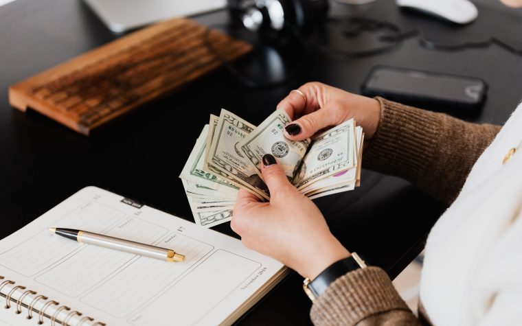 Unrecognizable elegant female in sweater counting dollar bills while sitting at wooden table with planner and pen Femme élégante non identifiable en pull comptant des billets de dollars assise à une table en bois avec un planificateur et un stylo