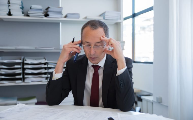 Businessman in a black suit stressed over documents in a bright office setting. Homme d'affaires en costume noir stressé sur des documents dans un environnement de bureau lumineux.